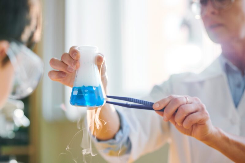 Hands of contemporary chemist or researcher holding glass tube with blue fuming fluid during experiment