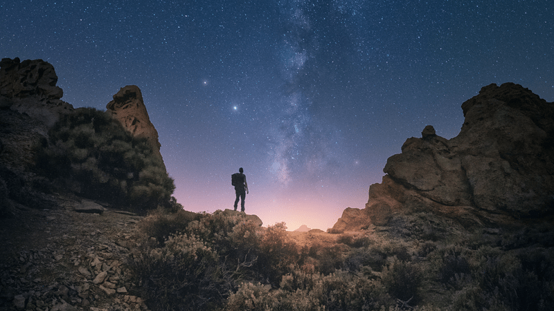 Man with backpack surrounded by mountains staring at a pink-purple sky