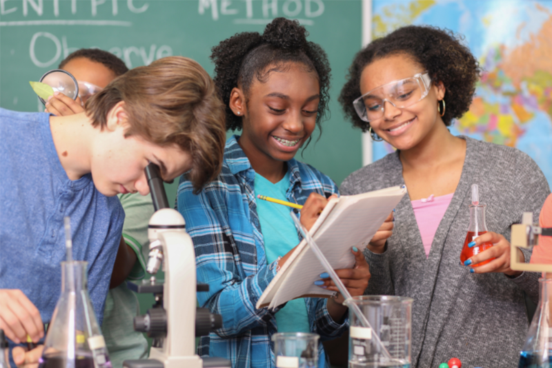 Three students looking into a microscope, writing on a pad of paper and holding a test tube