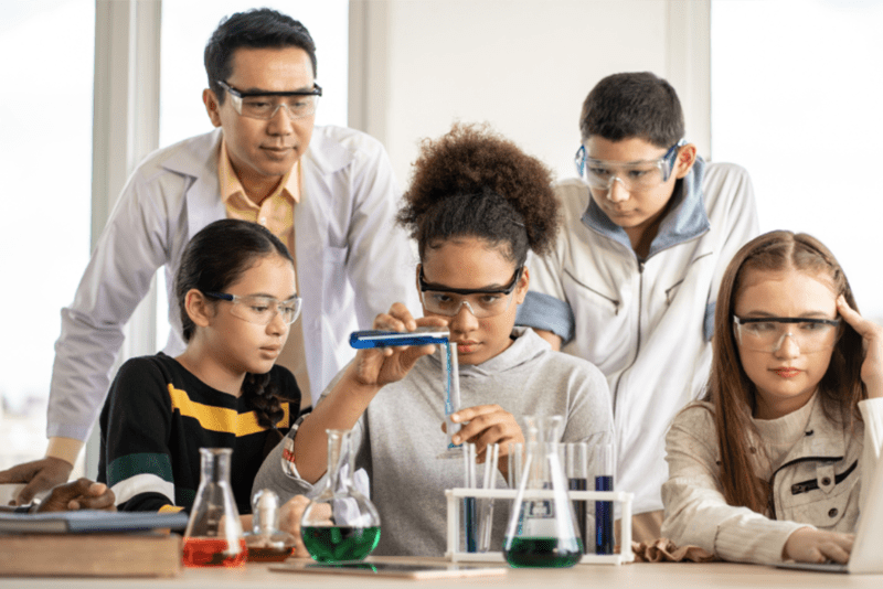 Three students and a teacher wearing safety goggles performing a science experiment with test tubes and liquids