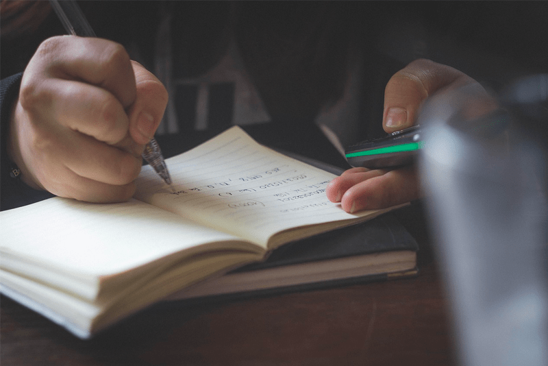 Student writing in a notebook with one hand and holding a calculator in the other